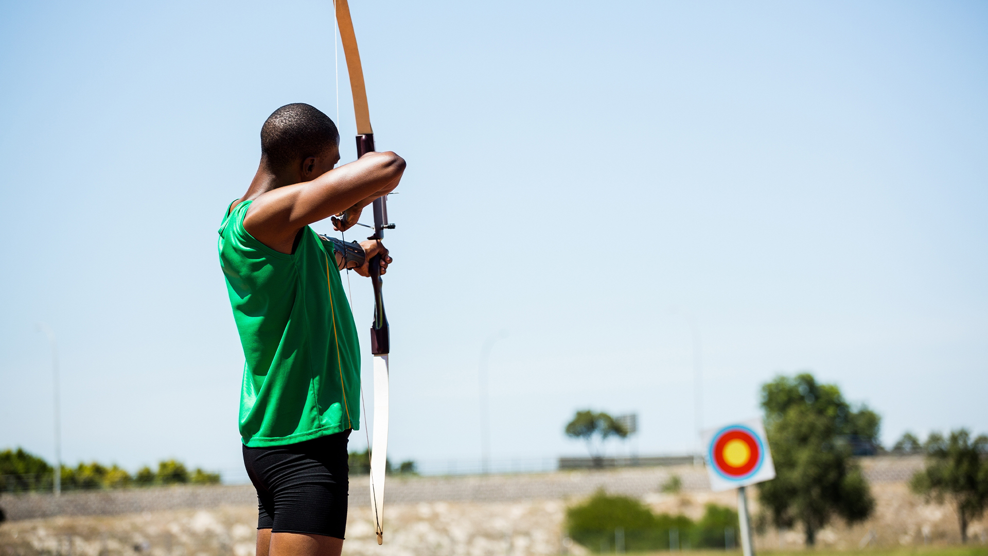Man stands with cross-bow aiming at a target in the distance. 